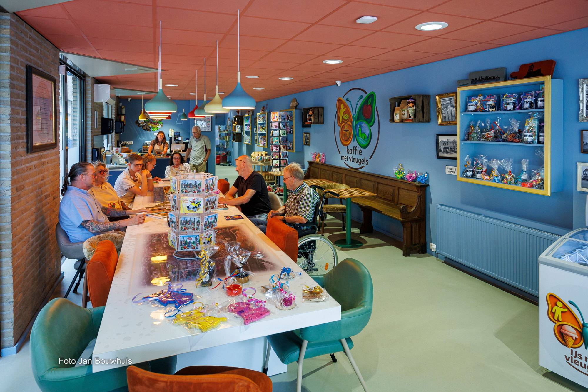 Overzicht van de koffiebar: de lange tafel met sleutelhangers en souvenirs, hanglampen, een kerkbank en de vitrine met kado's.