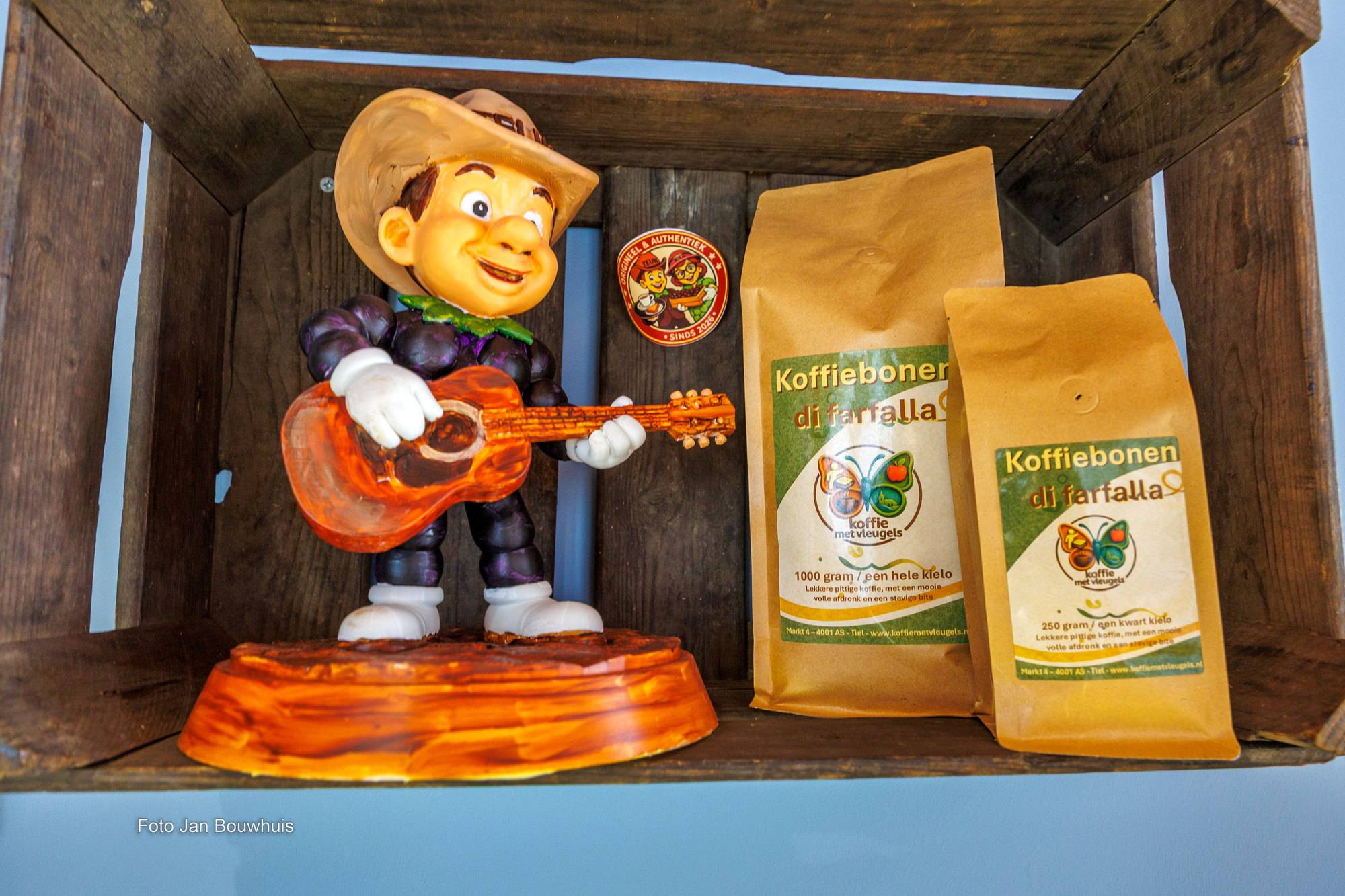 Een beeldje van een gitaarspeler in een houten kist, naast zakken koffiebonen di farfalla van Koffie met Vleugels.