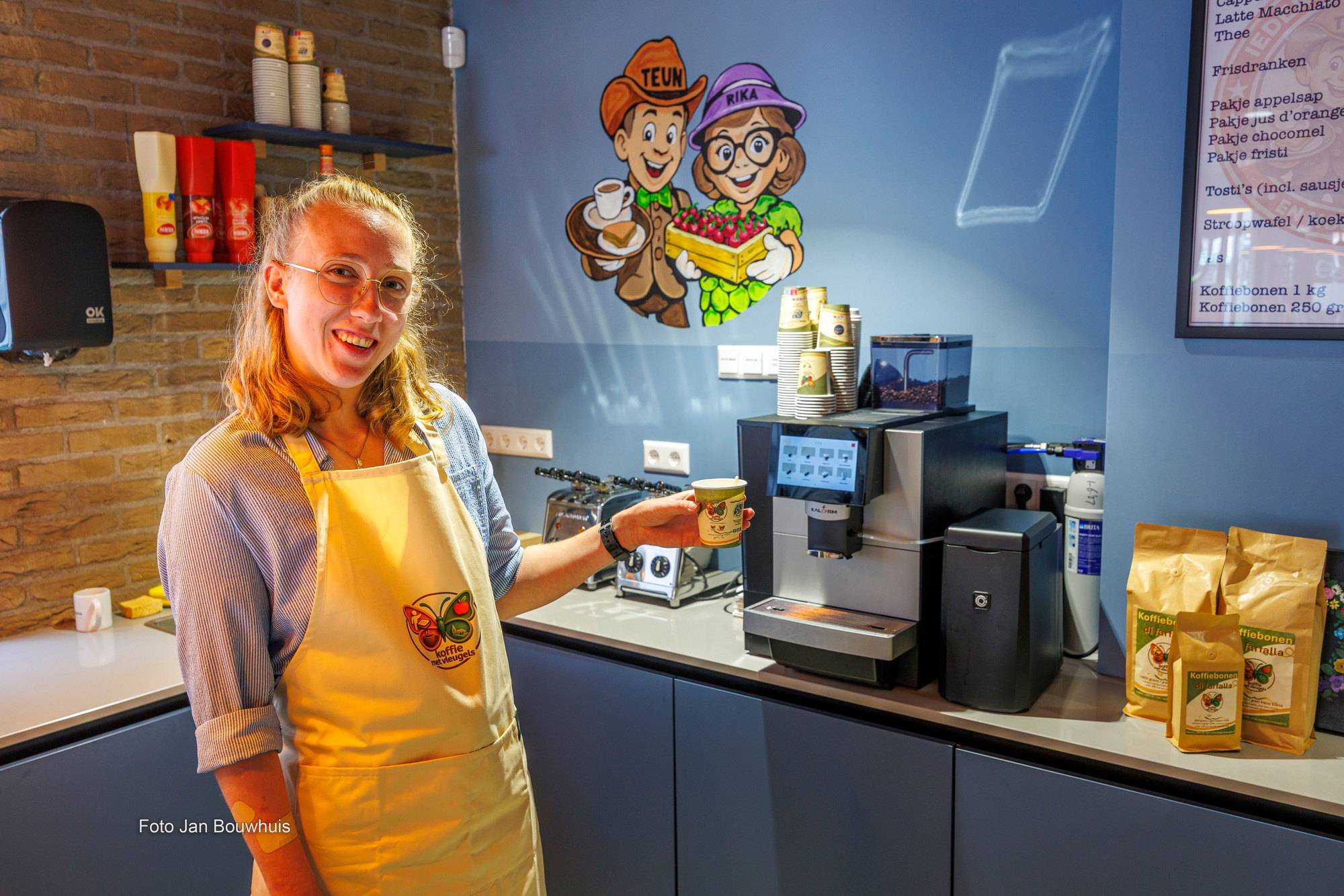 Een medewerkster in schort met een kop koffie bij de koffiemachine, met de tekening van Teun en Rika op de muur.
