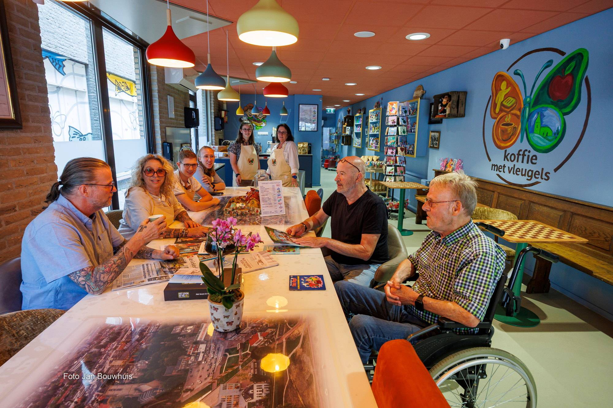 Bezoekers aan de lange tafel in de koffiebar, in gesprek met een kop koffie, met de vlinder van Koffie met Vleugels op de blauwe muur.