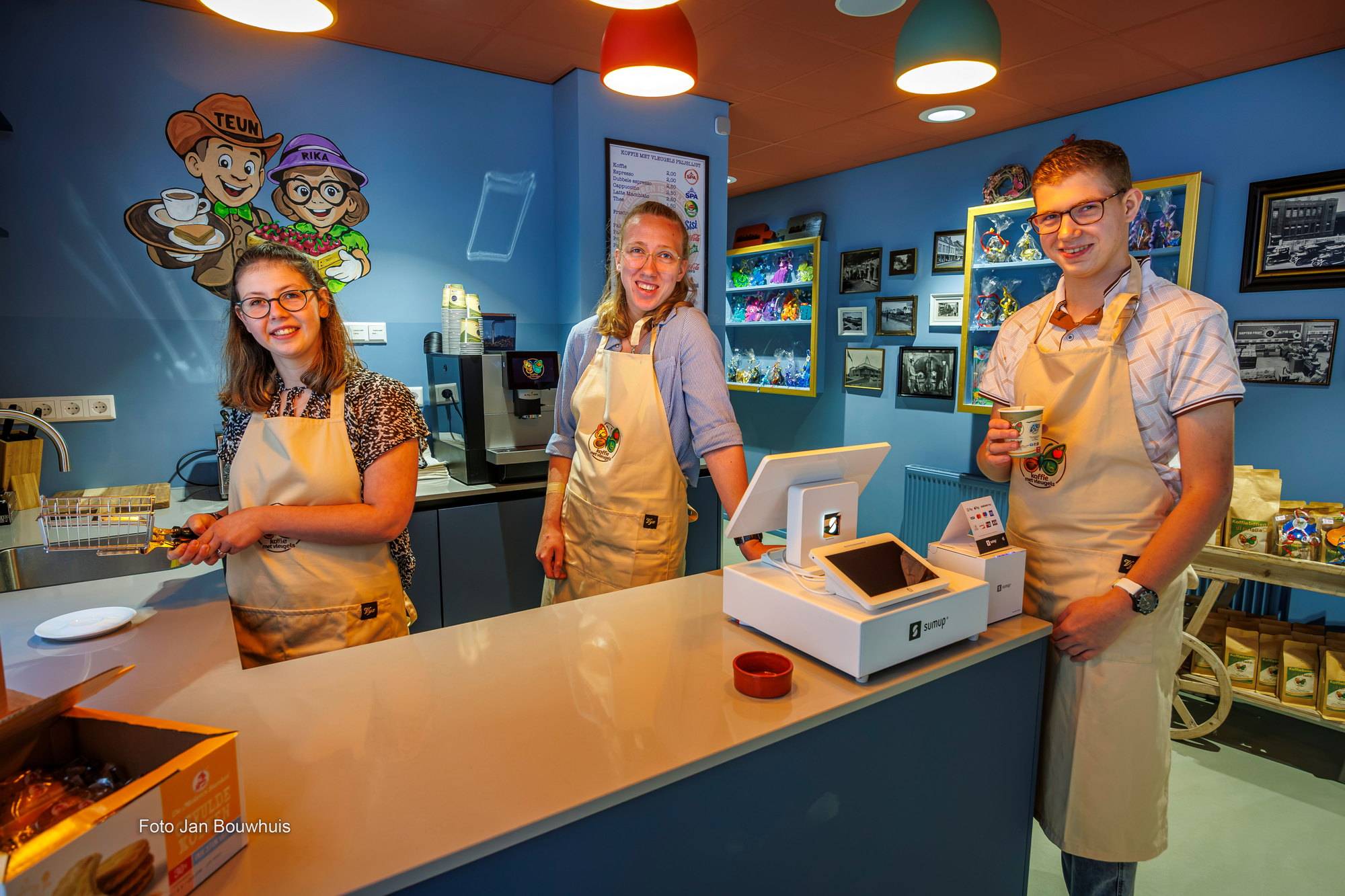 Drie medewerkers in schort achter de toonbank bij de kassa, met oude foto's en souvenirs aan de muur.