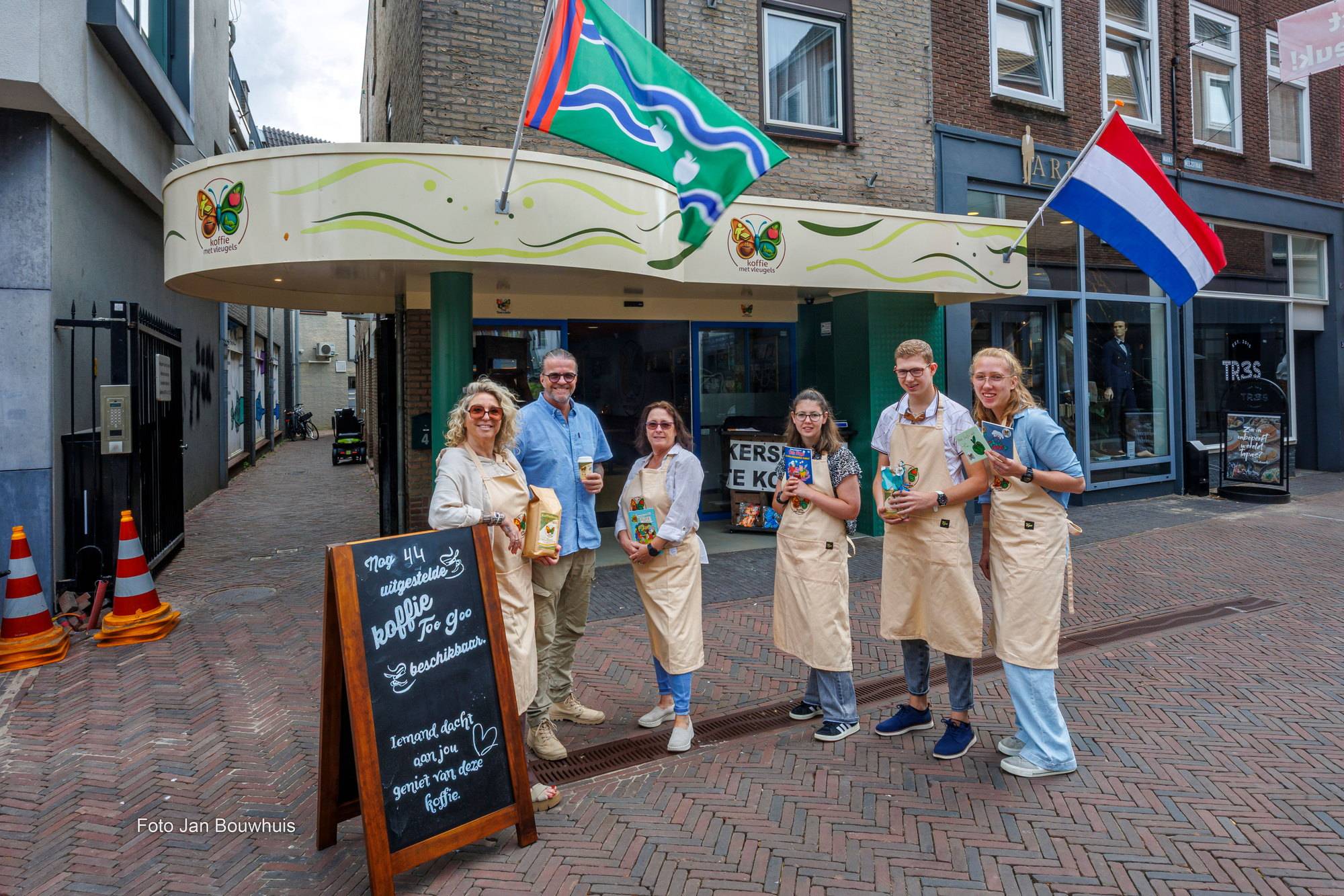 Het team van Koffie met Vleugels in schort voor de deur aan de Markt, naast het krijtbord.